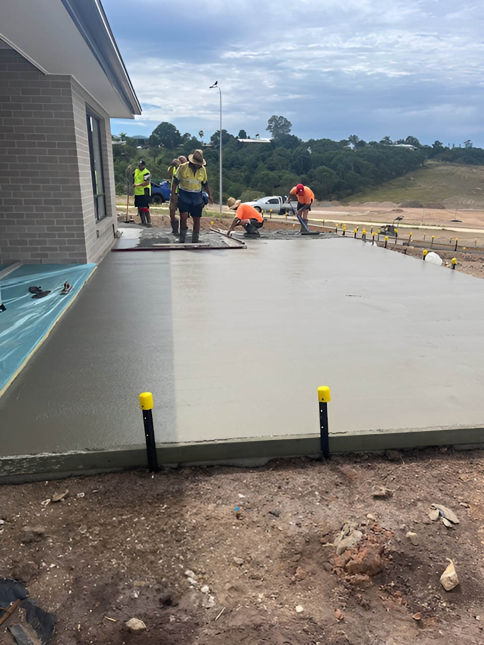 Concreters finishing a freshly poured slab beside a brick house, tall view