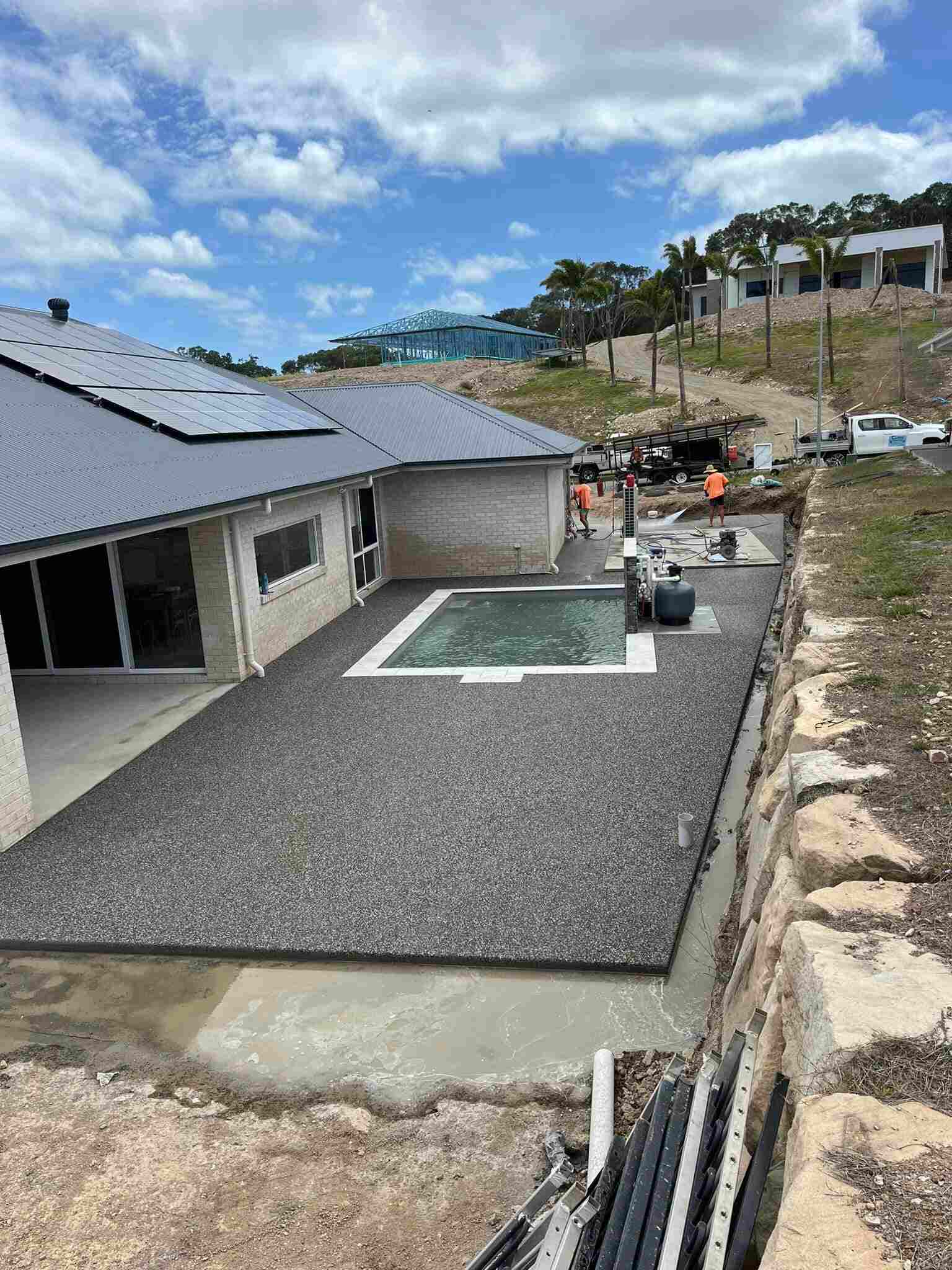 Exposed aggregate pool surround beside a brick home with workers in the background